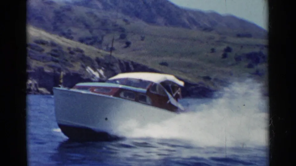 A mid-century motorboat races across a California lake, throwing a white wake against the hilly shoreline.