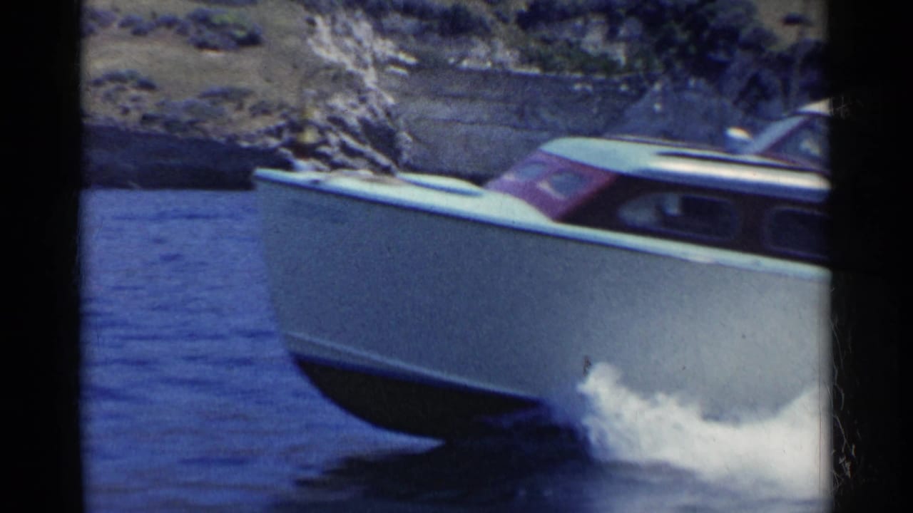 A white 1950s speedboat cuts across blue water in California, with spray at the bow and rugged shoreline behind it.