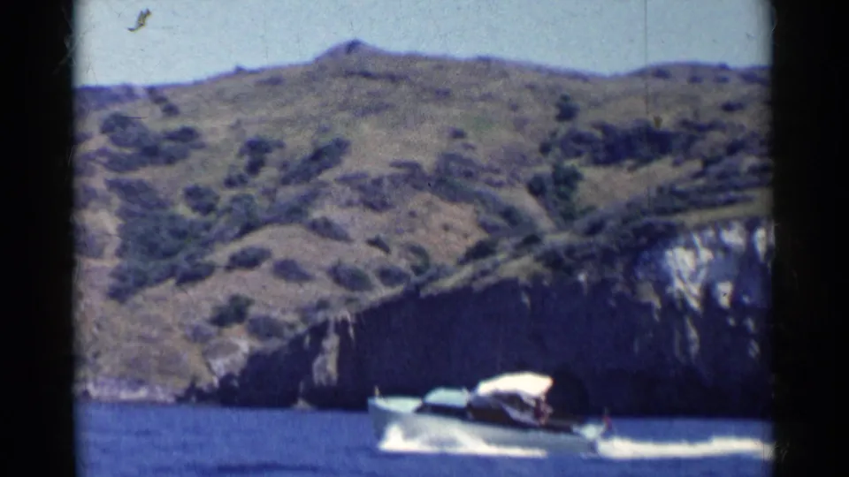 A white speedboat moves across blue water near rocky cliffs, leaving a broad wake in this California 8mm home-movie frame.