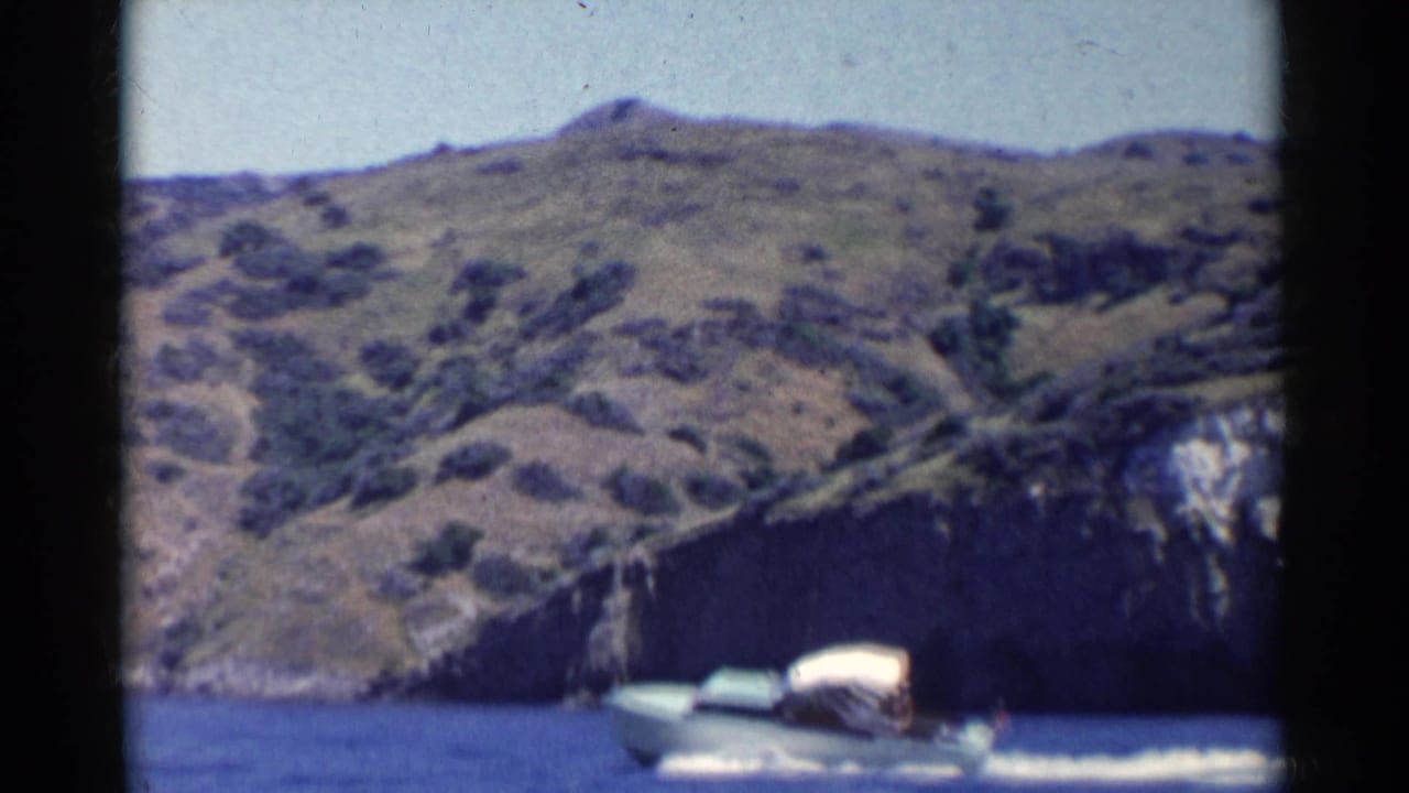 1955 California 8mm frame showing a small white canopy power boat passing below rugged brown-green hills and rocky shoreline.