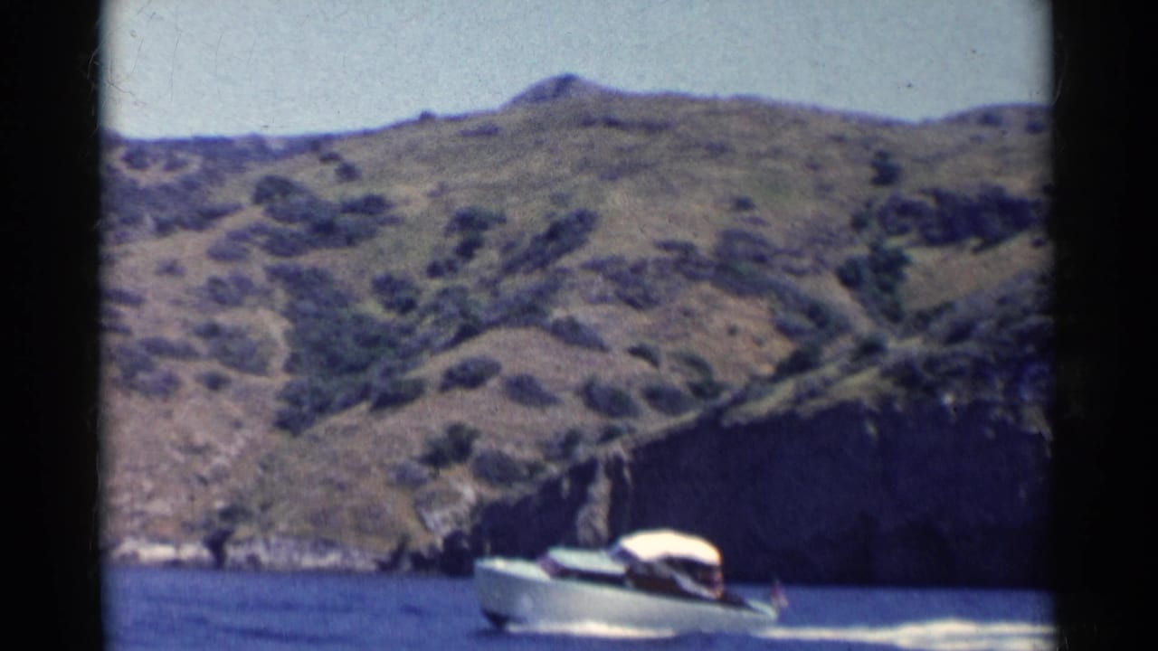 Canopy power boat passes near a rocky shoreline with steep hills behind it.
