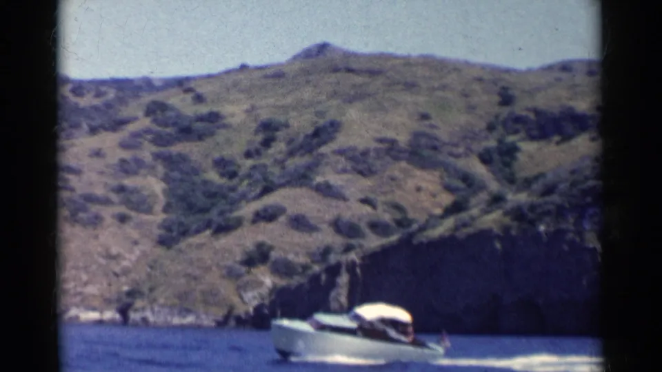 The white power boat continues along blue water in California, with a dark rocky cliff and sunlit chaparral hills in the background.
