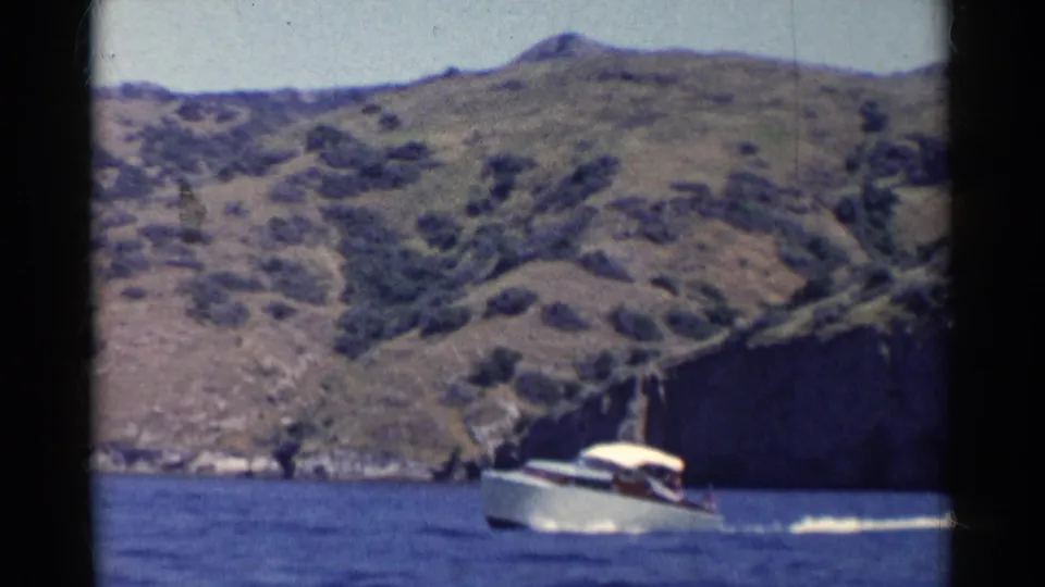 A small white canopy boat travels across blue water under steep California hills, with soft 8mm color and visible frame edges.