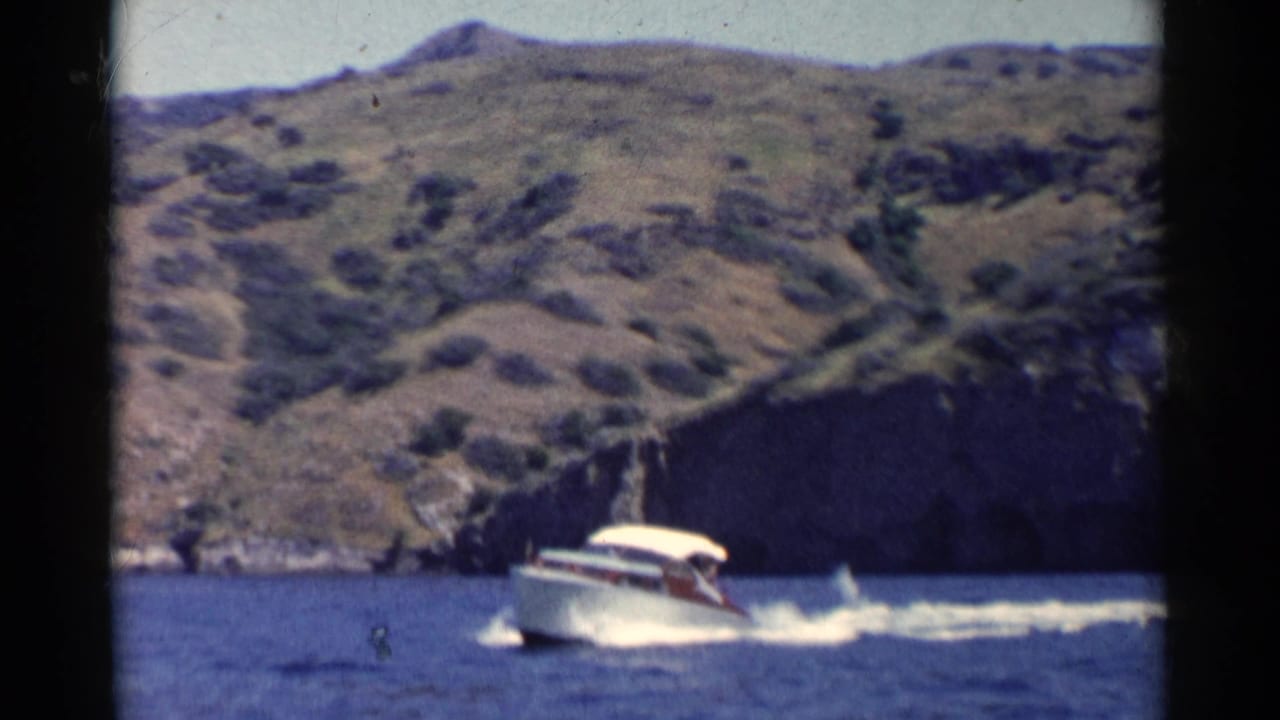 1955 California 8mm frame showing a white motorboat with canopy moving across blue water near dry coastal hills.