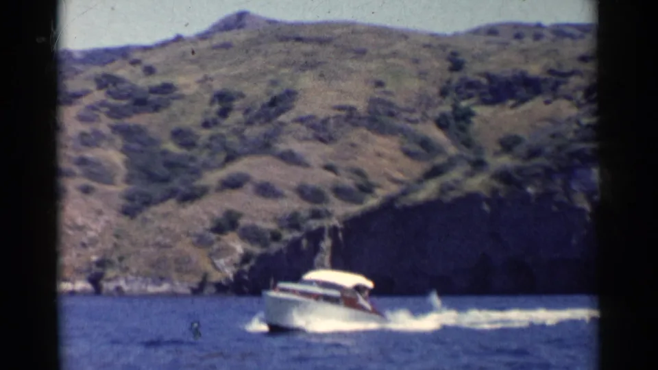 1955 California 8mm frame showing a white motorboat with canopy moving across blue water near dry coastal hills.
