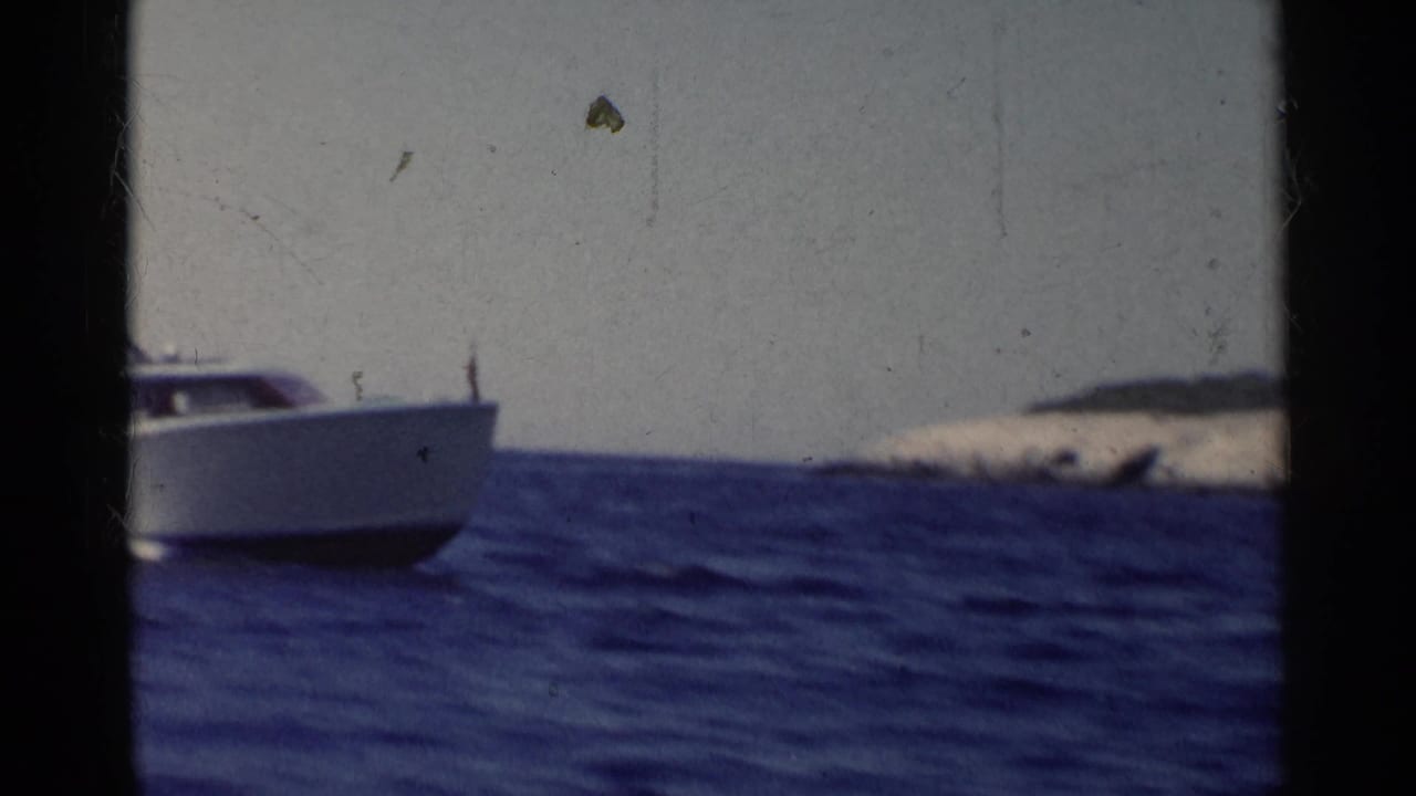 1955 California 8mm frame showing a white speedboat partly in view on deep blue water near a low rocky island.