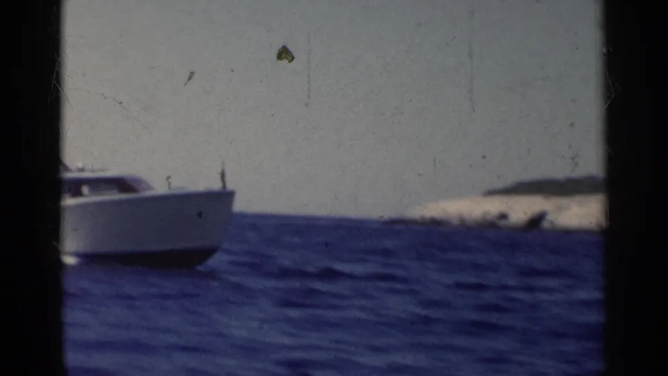 1955 California 8mm frame showing a white speedboat partly in view on deep blue water near a low rocky island.