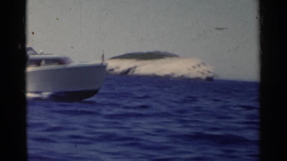 White speedboat moves across blue water in California home-movie footage, with a low rocky island under a pale sky.
