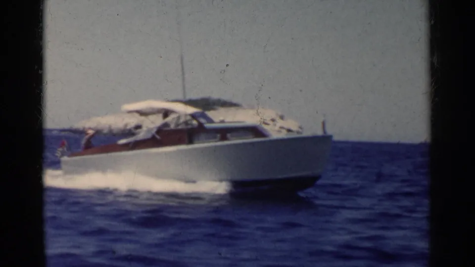 1955 California 8mm frame of a white cabin speedboat at speed, with spray and a rocky island visible behind it.