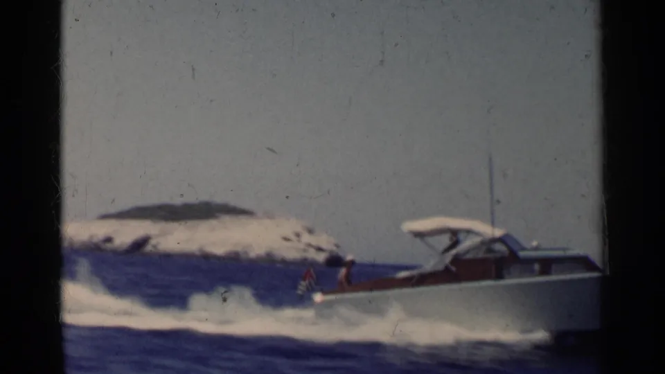 White speedboat leaves the frame in 1955 California 8mm footage, with wake spray, open water, and a rocky island in view.
