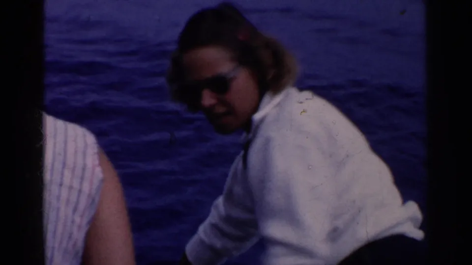 1955 California home-movie frame showing two adults aboard a boat, one in a white shirt and sunglasses, with ocean water behind them.