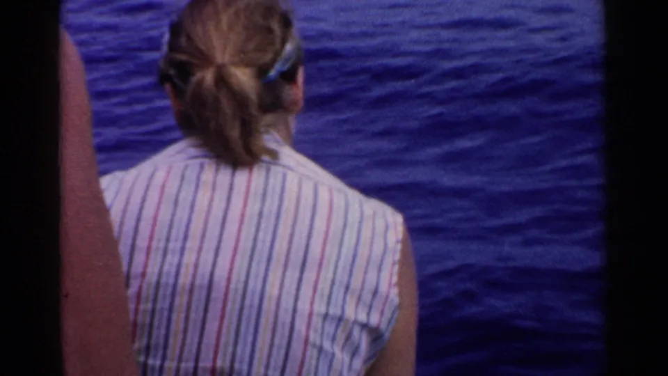 1955 California home-movie frame of a passenger viewed from behind on a boat, with rippled blue ocean water beyond.