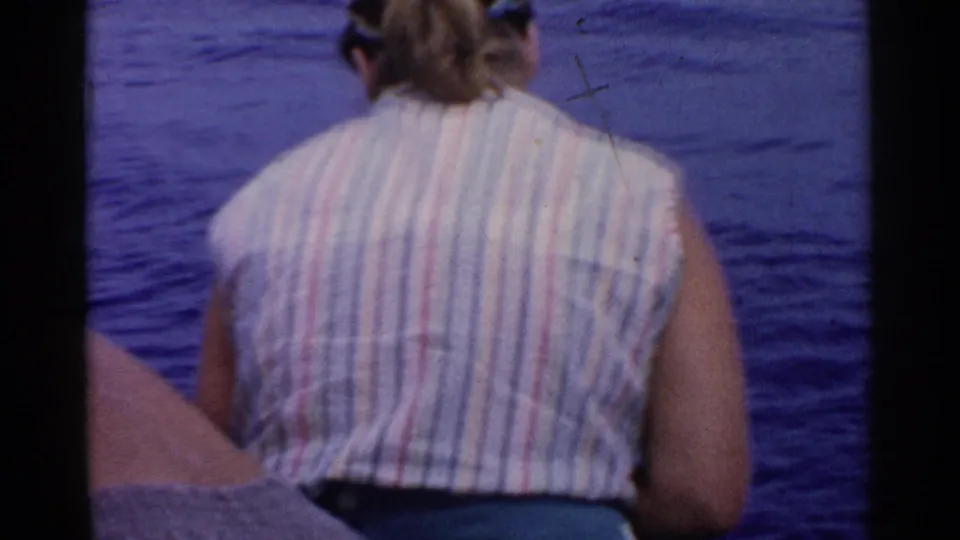 1955 California 8mm frame showing a seated passenger in a striped sleeveless shirt near the boat edge with ocean water behind.