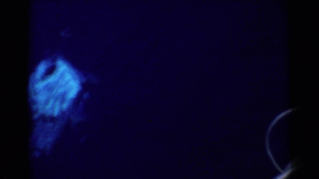 1955 California 8mm frame showing dark blue water and a faint pale shape below the surface, with heavy grain and edge darkness.