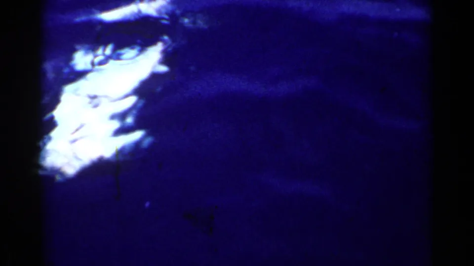 Dark blue water with a bright blurry white underwater form, possibly a shark or large fish, in the 1955 California home-movie footage.