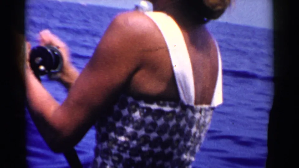 In 1955 California home-movie footage, a woman in a patterned swimsuit with white straps holds a fishing rod on a boat at sea.