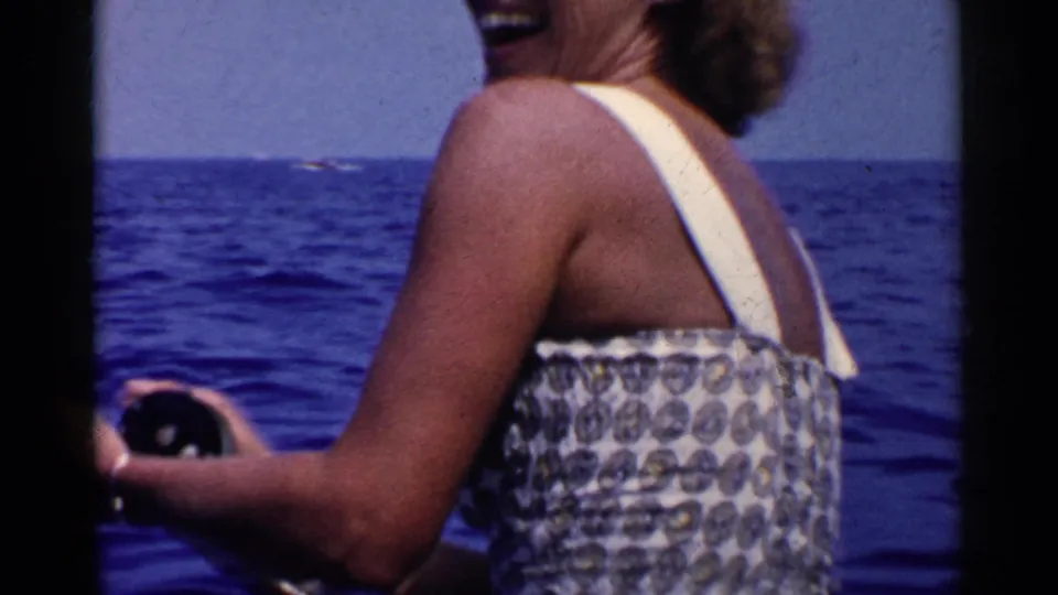 The woman smiles while holding fishing gear on a boat, with deep blue ocean and a distant horizon visible behind her.