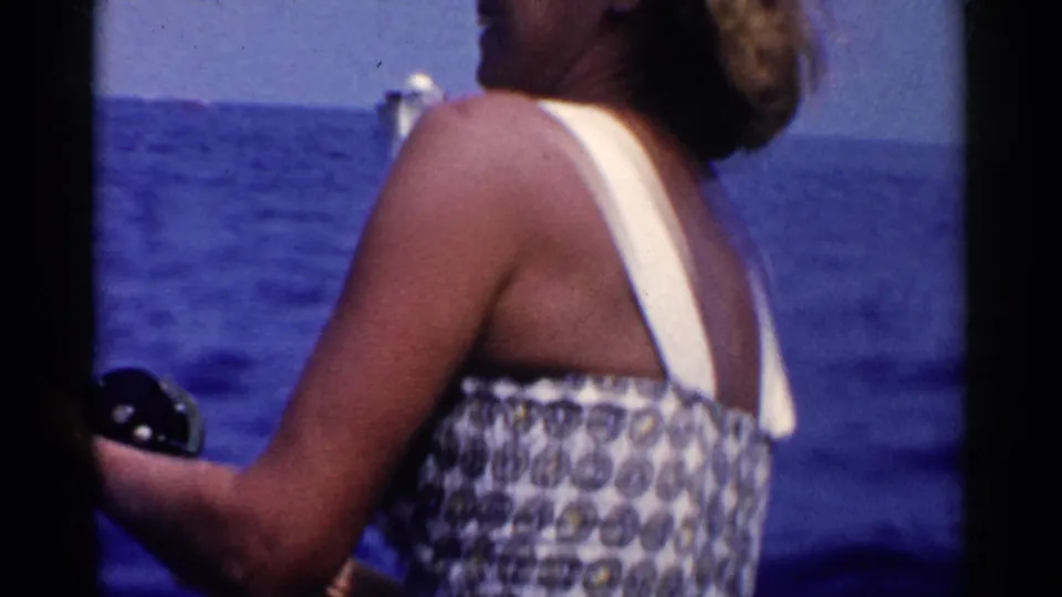A side view shows the woman in a patterned swimsuit holding fishing equipment, with open ocean and a small distant boat behind her.