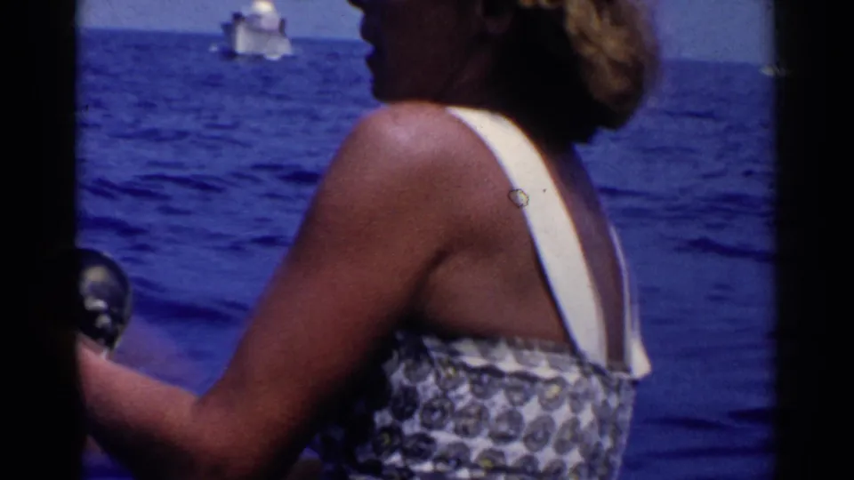 The woman stands with her back partly turned while fishing or looking out from the boat, with a distant vessel on the blue ocean.