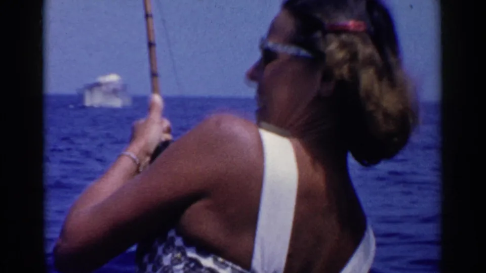 A woman in a patterned swimsuit holds a fishing rod on California water, with a small vessel visible in the distance.