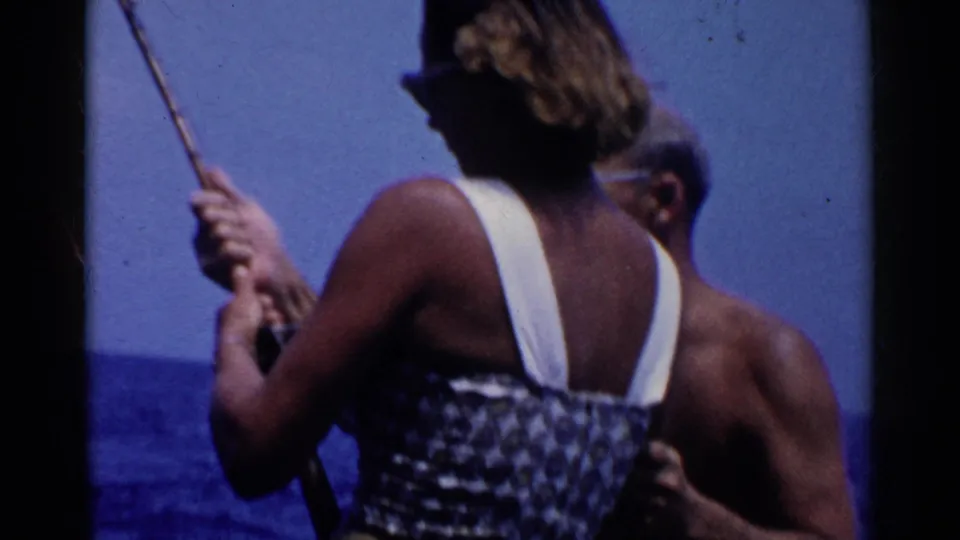 The woman continues fishing from the boat as a shirtless man stands close behind her in the 1955 California water scene.