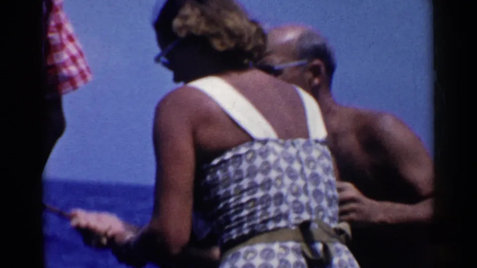 The woman and a shirtless man stand together on a boat in supported 1955 California footage, with blue water and sky behind them.