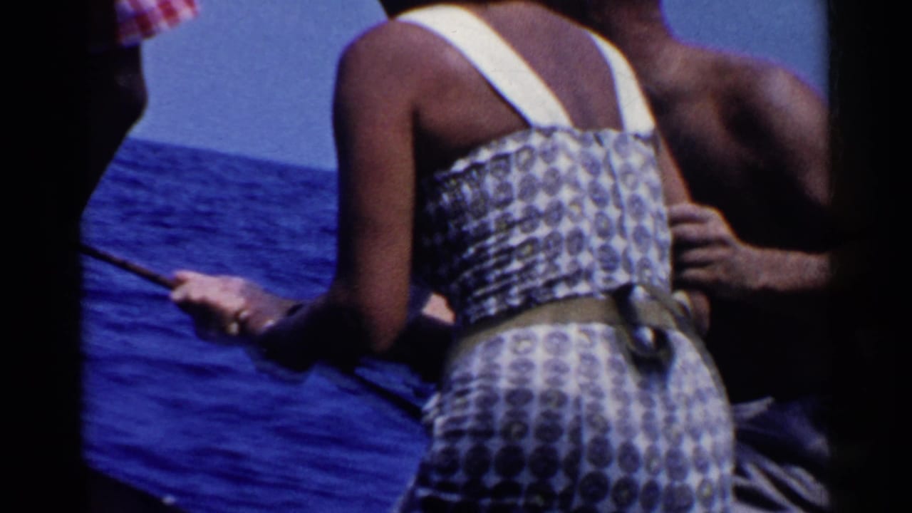 Woman in a patterned sleeveless dress holds a fishing rod on a boat with ocean behind her.