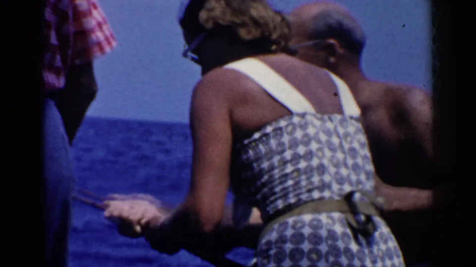 A woman in a white-strap patterned dress stands with a fishing rod near a shirtless man in 1955 California boating footage.