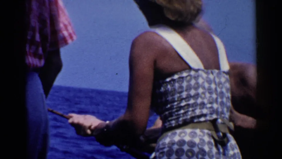 1955 California home-movie frame of a woman in a patterned summer dress on a boat as a man in plaid stands nearby over blue ocean.