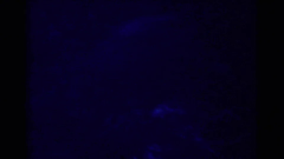 Dark underwater 8mm frame from the fishing sequence, with faint fish-like shapes moving through deep blue water.