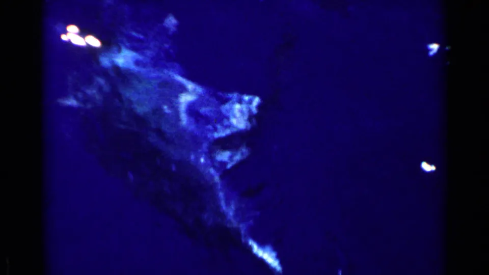 1955 California 8mm frame showing a pale irregular form in deep blue water, possibly marine life or rock seen through glass, with small bright reflections nearby.