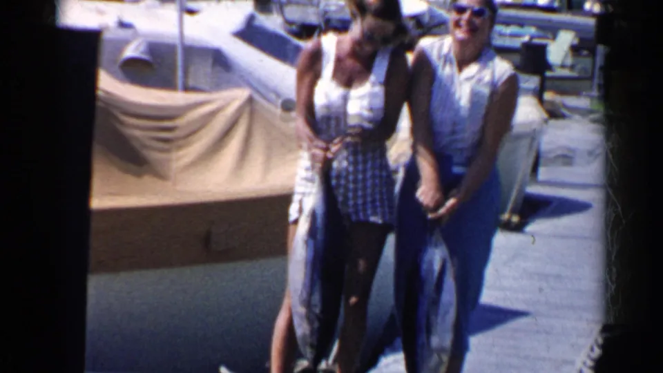Two adult women in 1950s-style summer clothing stand on a dock in California, holding large fish beside a boat in bright sun.