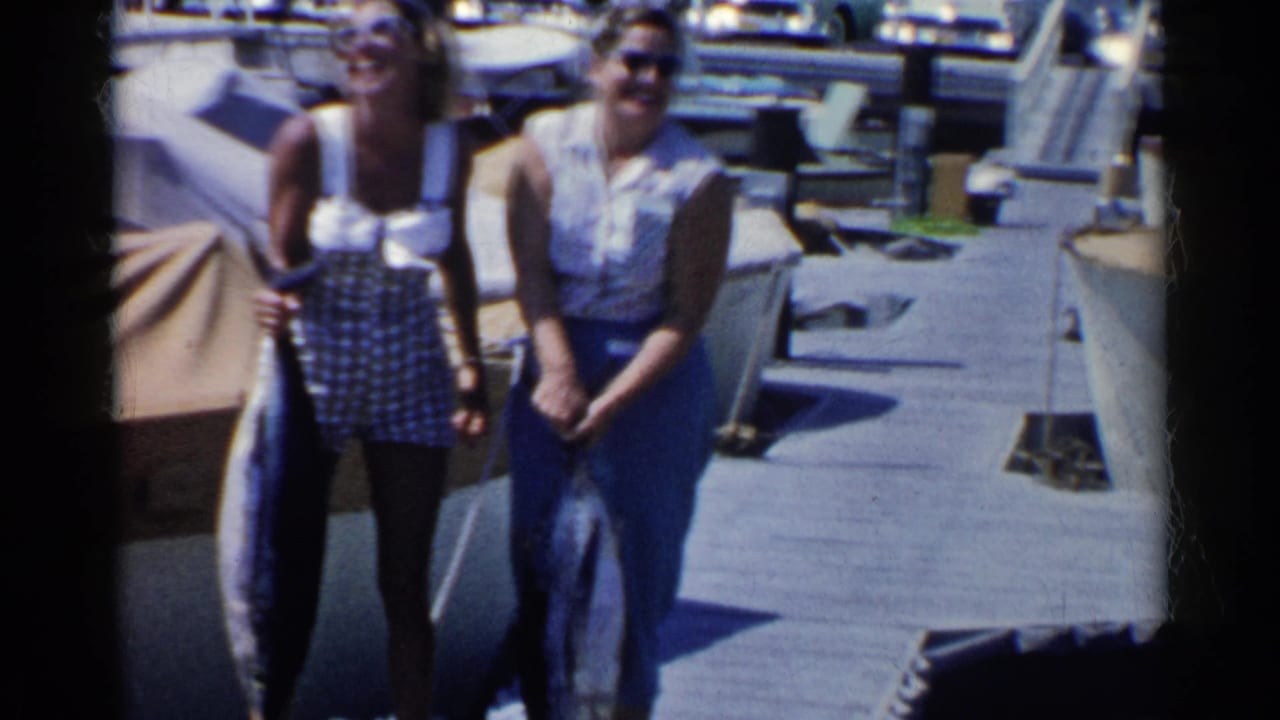 Two women standing on a dock while holding large fish near boats.