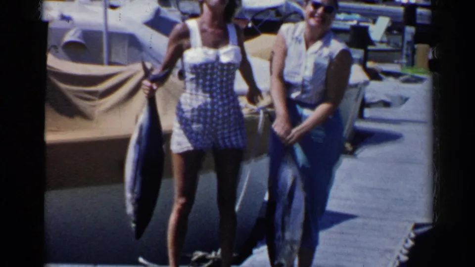 Two women in summer clothing smile toward the camera on a California marina dock while holding large tuna fish.