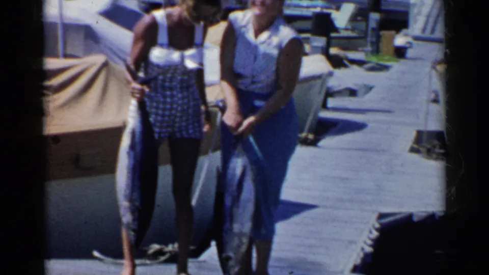 A closer 8mm frame shows two women on the dock with large fish hanging in front of them near marina boats.