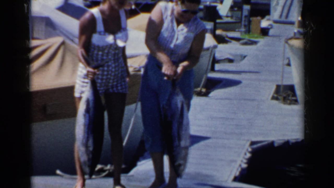 Two women on a sunny dock hold large fish beside moored boats.