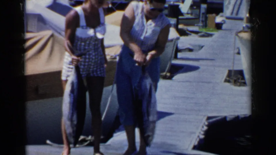 Two women in 1950s swimwear stand on a California marina dock holding large fish in bright sun.
