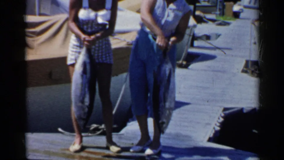 Two women on a California marina dock display large fish, with boats and dock fittings behind them.