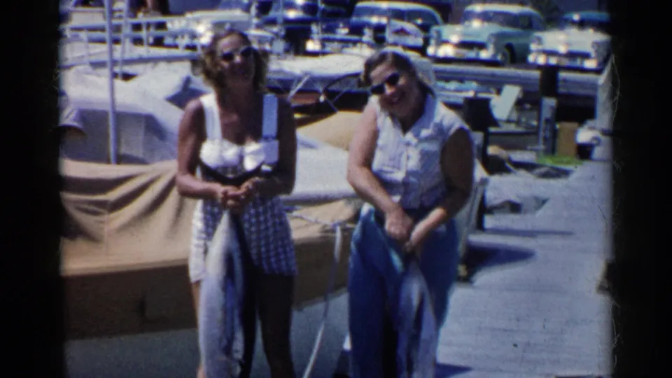 Two women in sunglasses and midcentury swimwear pose with fish on a sunny California marina dock, with cars in the background.