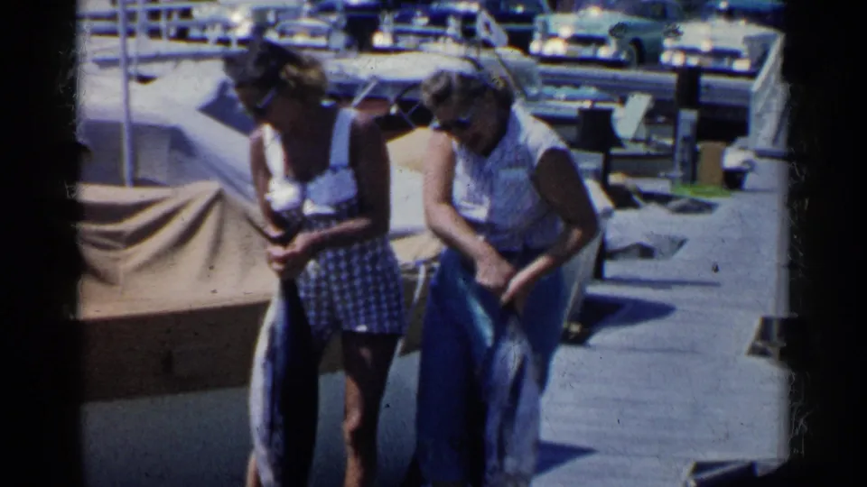 On a bright California marina dock, two women in 1950s swimwear handle large fish with boats and parked cars behind them.