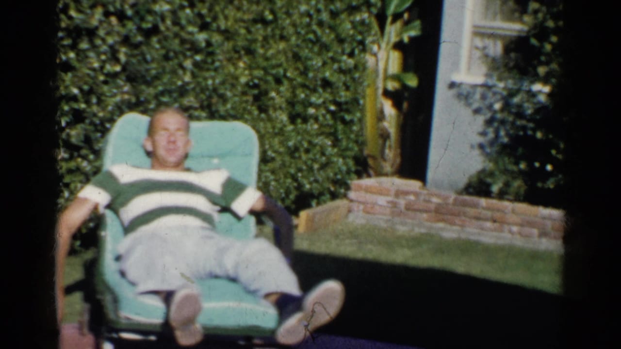 Man reclines in a turquoise lawn chair beside hedges and brick edging.