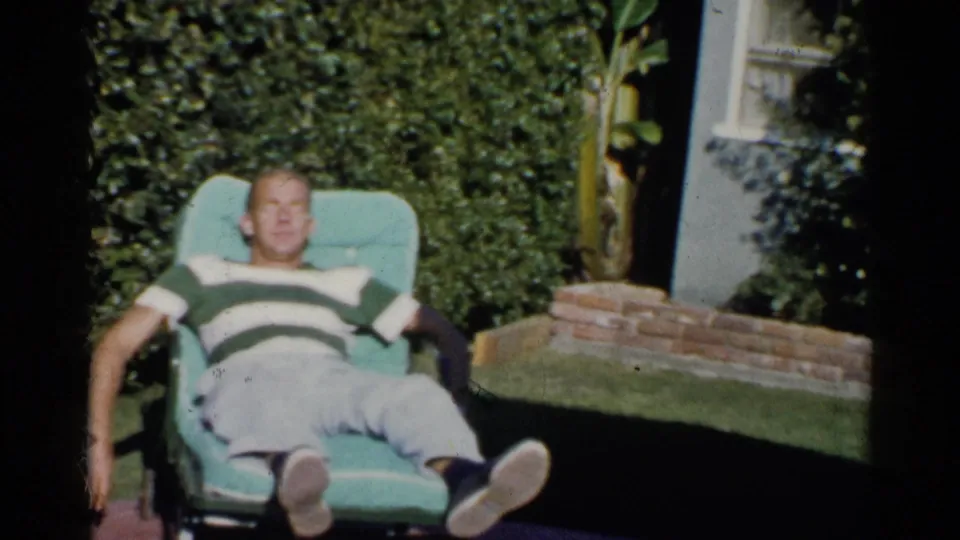 The man remains stretched out in a turquoise chair, framed by hedges, lawn, brick edging, and part of a house exterior.