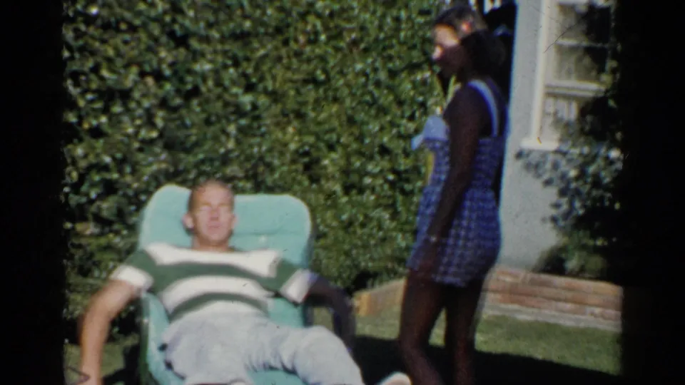 A woman in a blue checked dress approaches the reclining man in the sunlit California backyard scene.