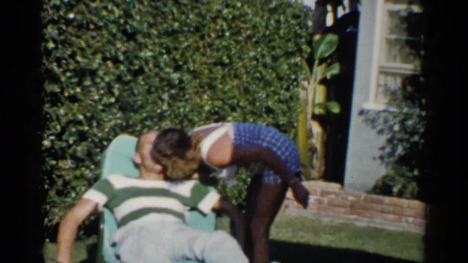 The woman bends toward the seated man and appears to kiss him on the cheek in a 1955 California backyard.
