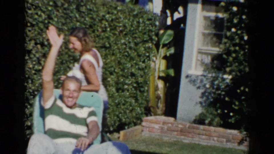 A smiling man in a striped shirt waves from a lawn chair as a woman passes behind him in a California backyard.
