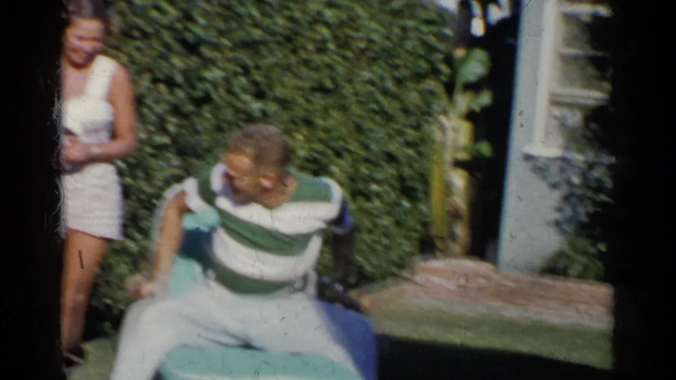 The woman stands at the side of the frame while the seated man turns in a lawn chair in the sunny California yard.