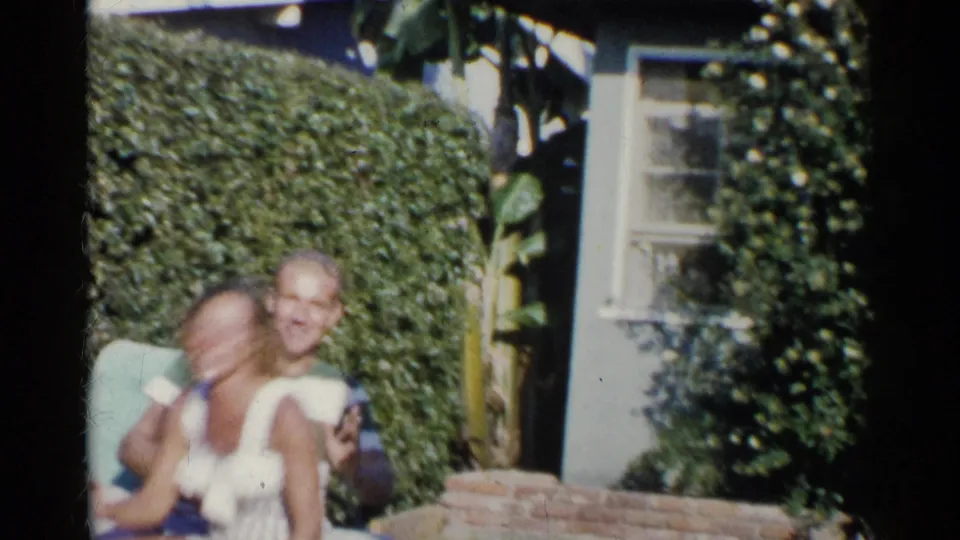 A woman laughs close to the camera as the seated man smiles behind her in the 1955 California backyard scene.