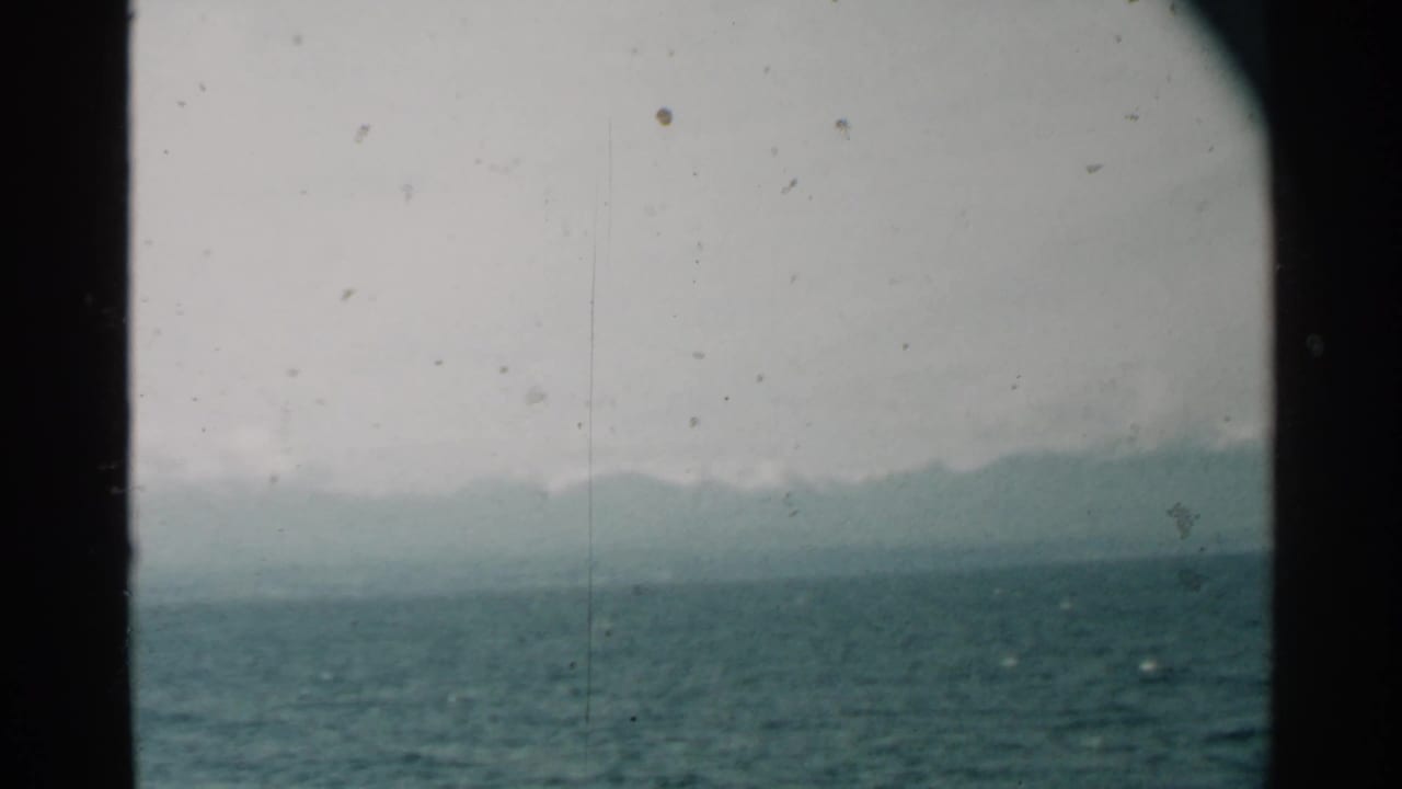 Porthole-framed view of choppy ocean with hazy mountains under a gray sky.