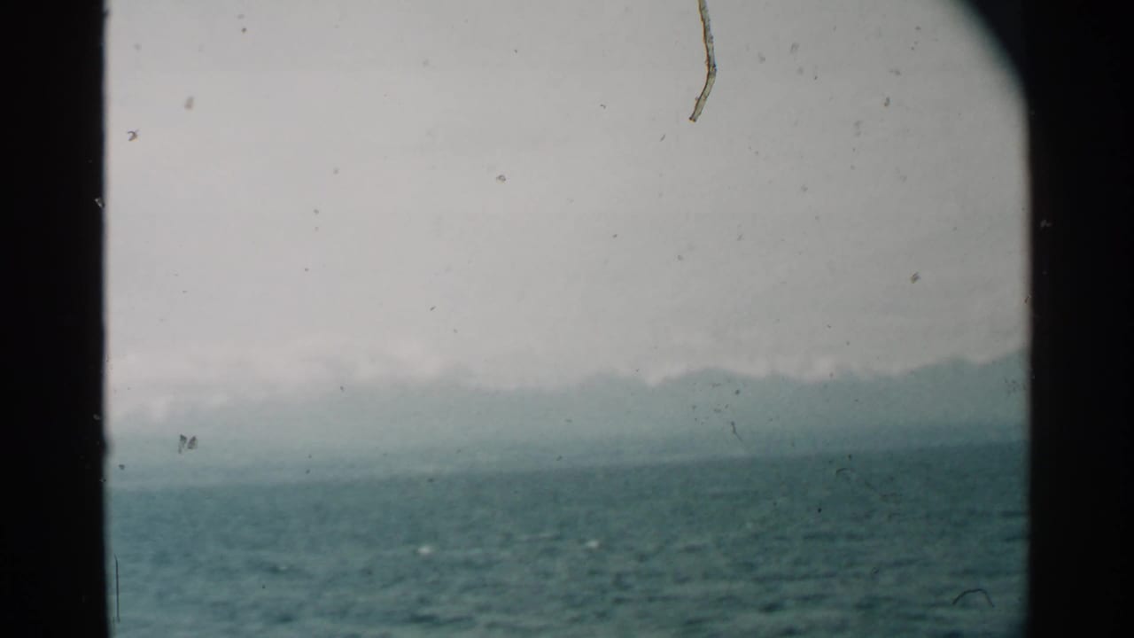 1958 Pearl Harbor view across textured water toward a hazy Hawaiian shoreline, divided by dark porthole framing.