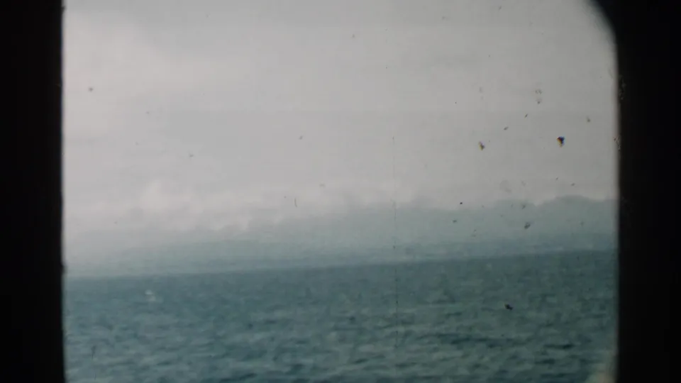 Mid-century 8mm frame from Pearl Harbor showing choppy water, low clouds, and a faint mountain line through porthole framing.